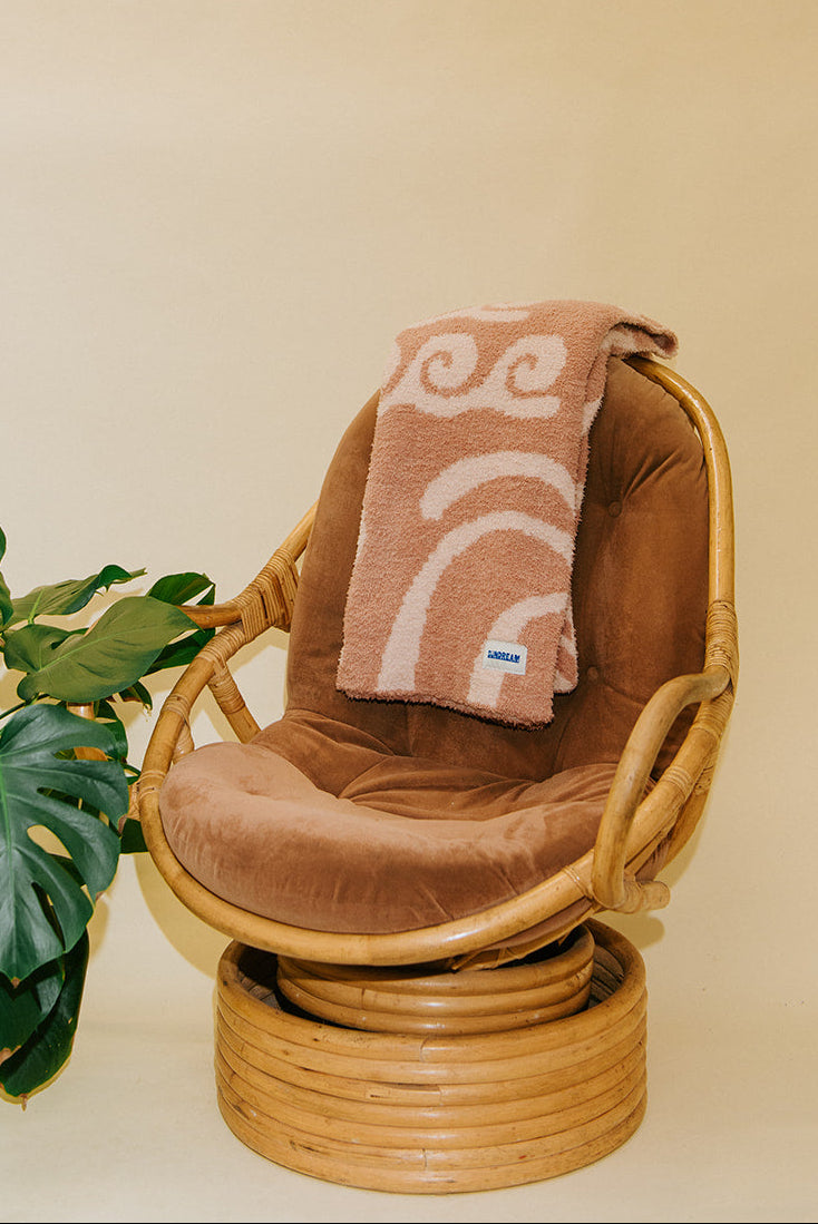 A terracotta-colored plush armchair with a matching cushion and a folded terracotta towel featuring a cream-colored wave pattern, styled next to a green plant against a warm beige background.