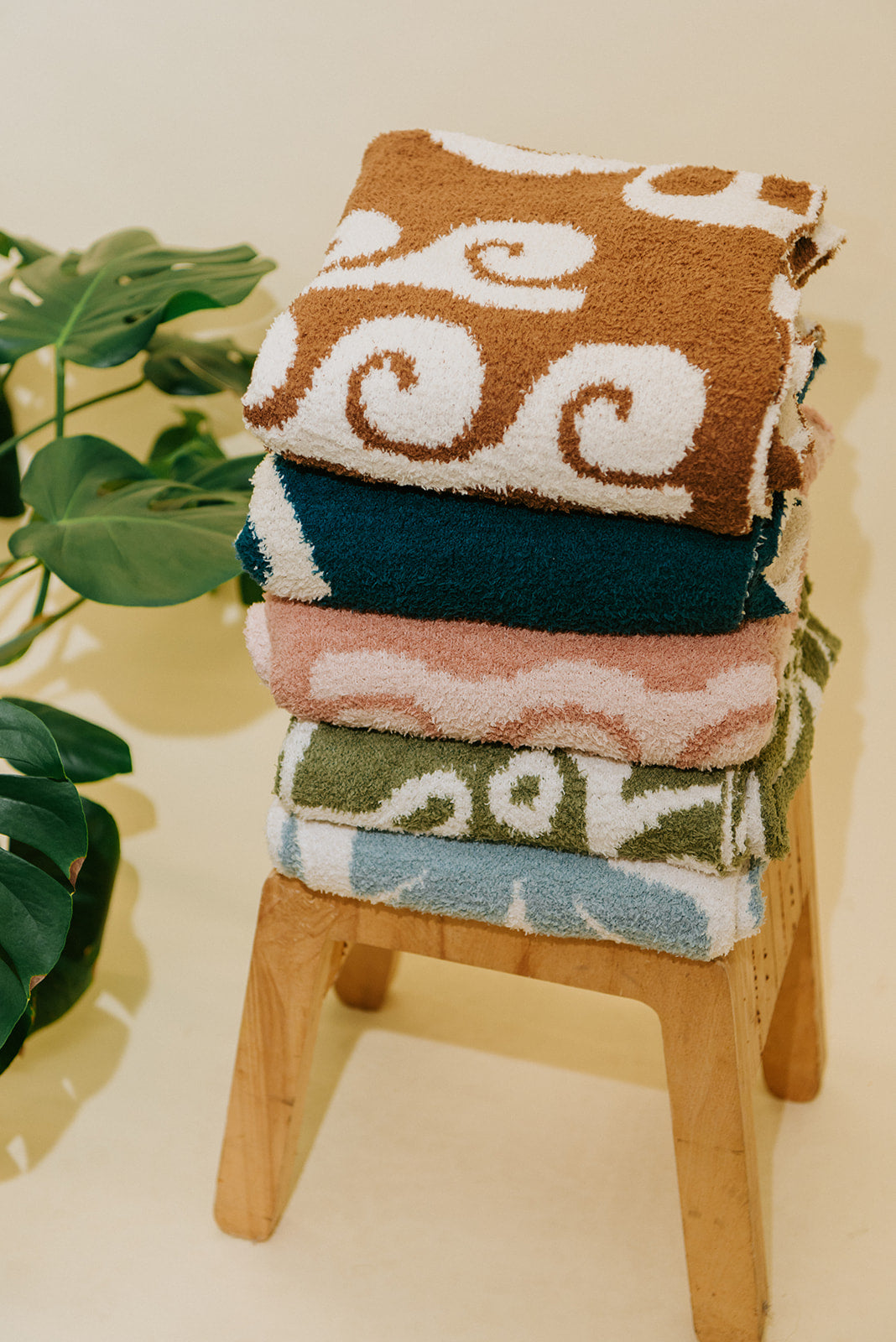 A stack of five folded patterned blankets displayed on a small wooden stool, with green plant leaves visible on the left against a warm cream background.