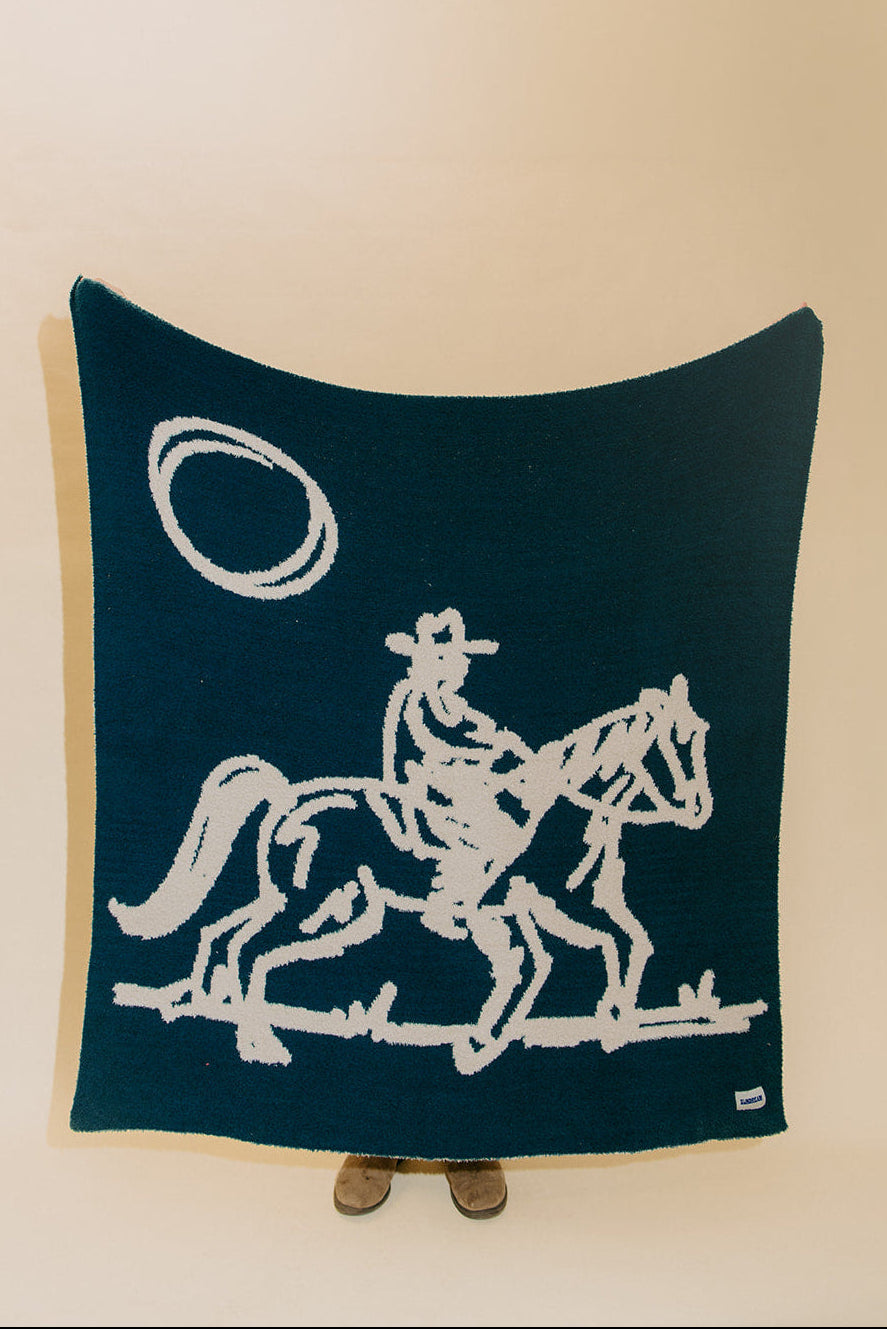 A dark teal throw blanket featuring a white hand-knit graphic of a cowboy riding a horse under a lasso, displayed against a plain beige background.