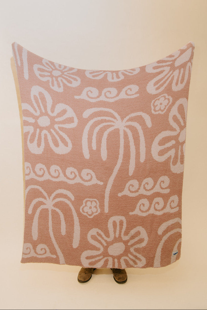 A rectangular throw blanket with a dusty pink background and a white hand-drawn style floral and palm tree pattern, displayed on a stand against a plain cream background.