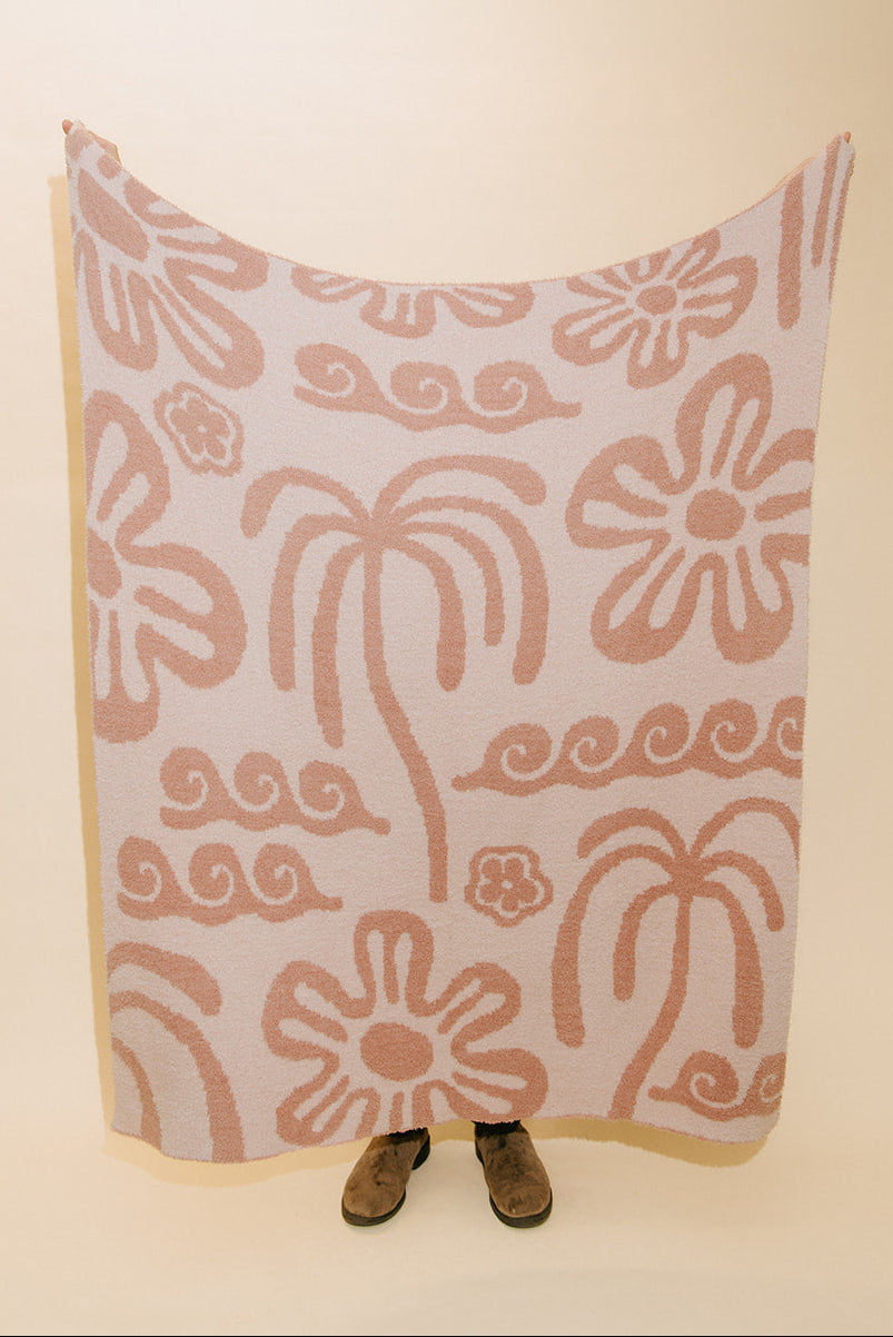 A large rectangular textile panel or blanket with a pink palm tree and floral pattern on a cream background, held up against a plain beige backdrop. Small brown shoes are visible at the bottom edge.