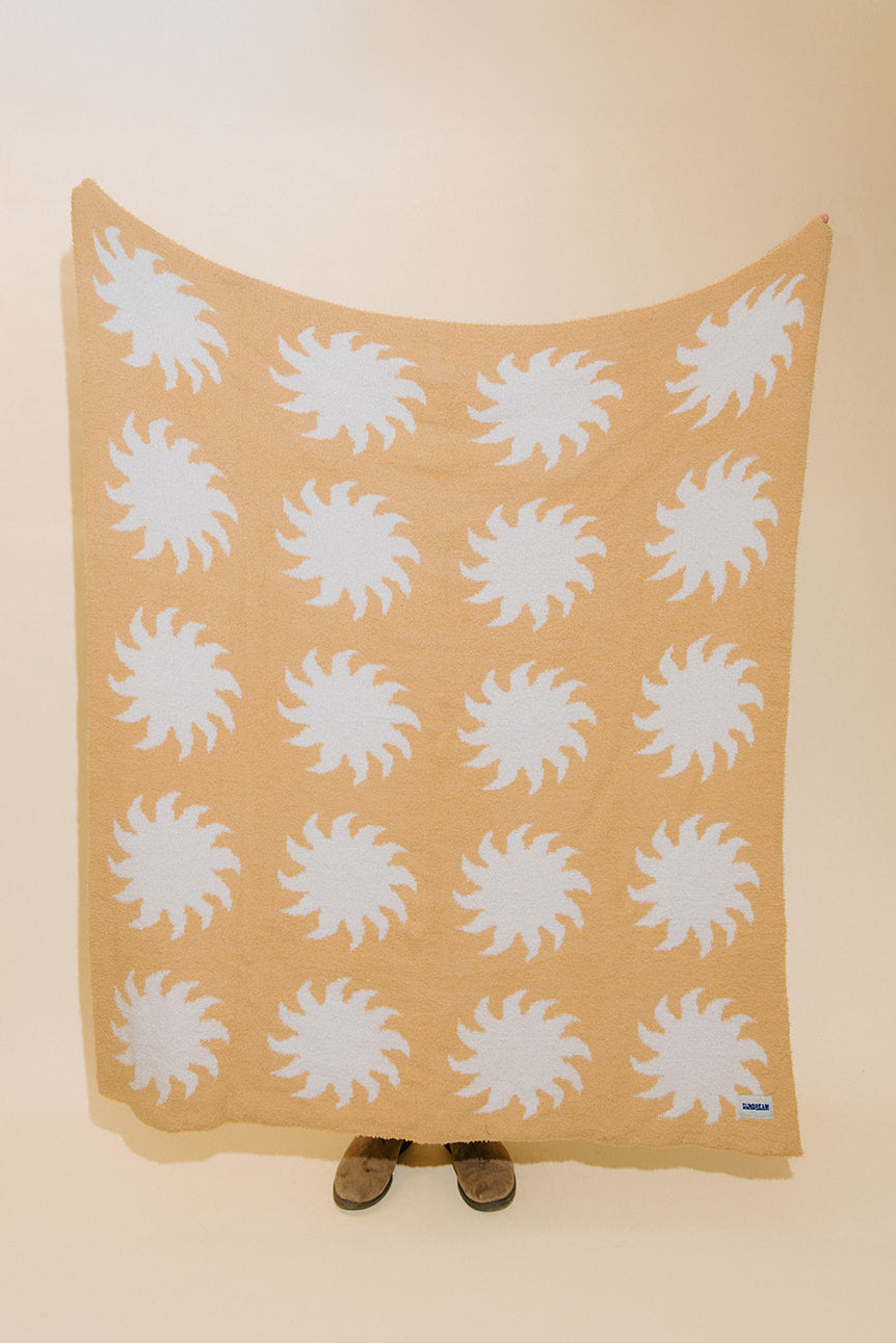 A beige blanket or throw with a repeating white sunburst pattern, held up against a plain beige background. Small brown shoes are visible at the bottom edge.