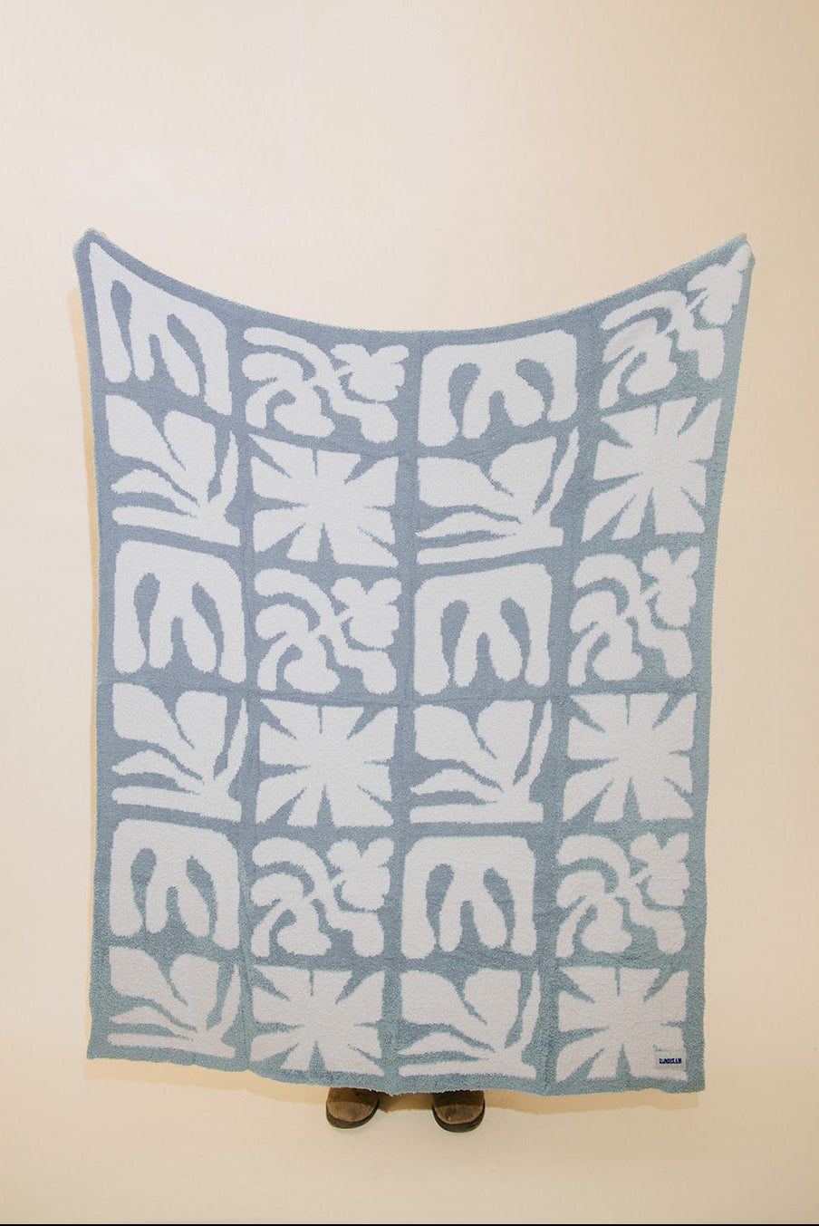 A light blue and white patterned blanket or throw is draped over a stand against a plain beige wall. The blanket features a repeating grid of abstract floral and leaf-like motifs.