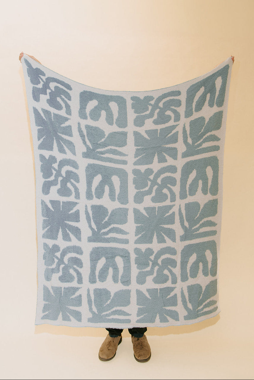 A person holds up a large square textile with a repeating abstract floral or leaf-like pattern in muted blue-gray on a white background, photographed against a plain warm off-white wall. The person's hands and feet are visible at the edges and bottom.