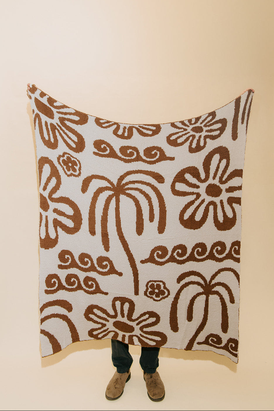 A person stands holding up a large blanket or throw with a brown abstract floral and swirl pattern on an off-white background, with their legs and shoes visible underneath.