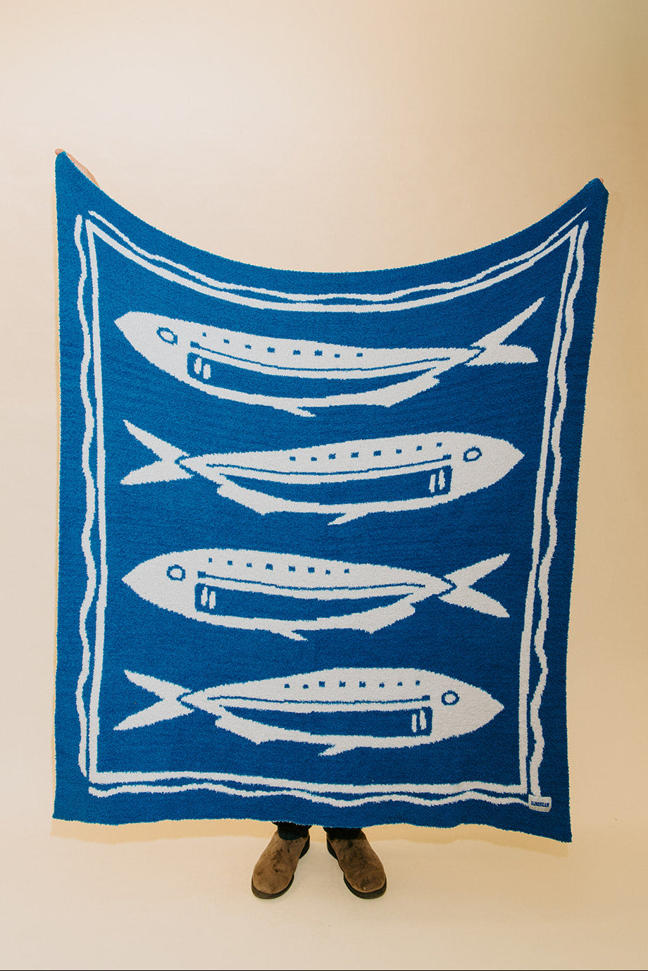 A blue blanket or throw featuring four white fish illustrations, held up by a person whose feet are visible at the bottom against a plain beige background.