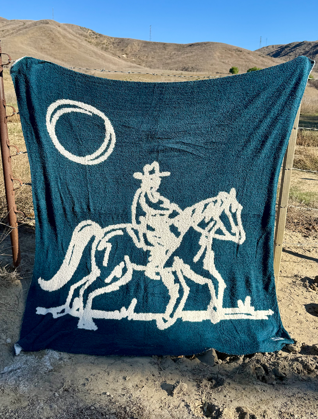 Navy blue blanket with white cowboy and horse design draped over a fence in a desert landscape.