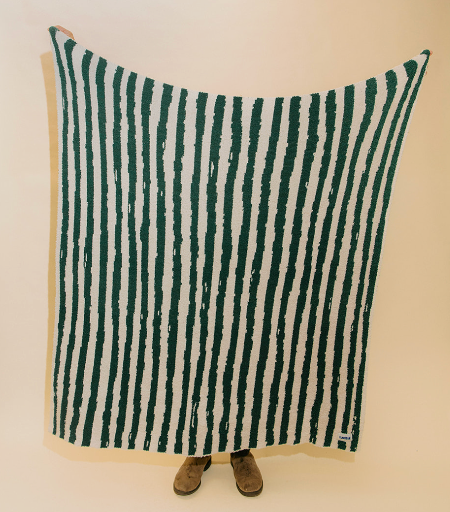 A large throw blanket with vertical dark green and off-white stripes is held up by a person whose feet are visible at the bottom, against a plain beige background.