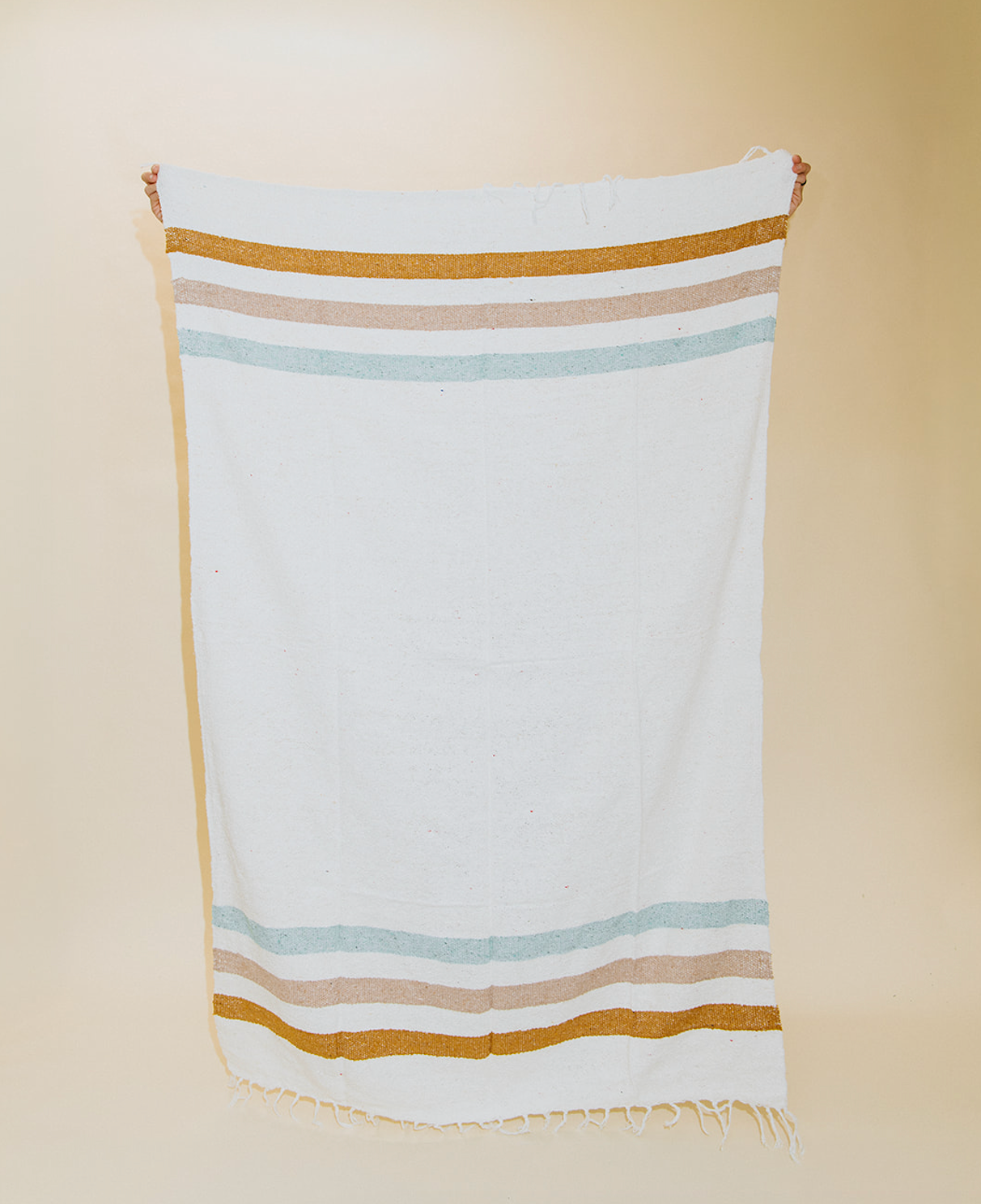 A white woven textile towel or scarf with horizontal stripes in tan, light blue, and beige, hanging against a plain beige background.