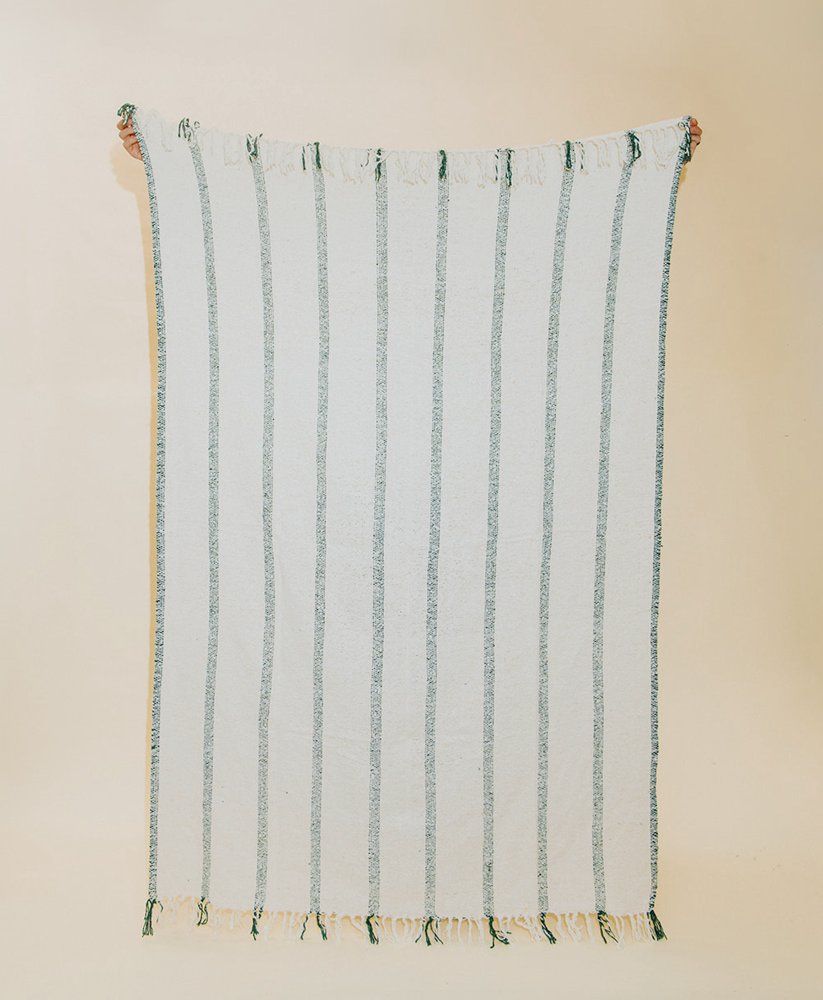 A white woven textile with vertical sage green stripes and fringed edges, hanging against a plain beige wall.