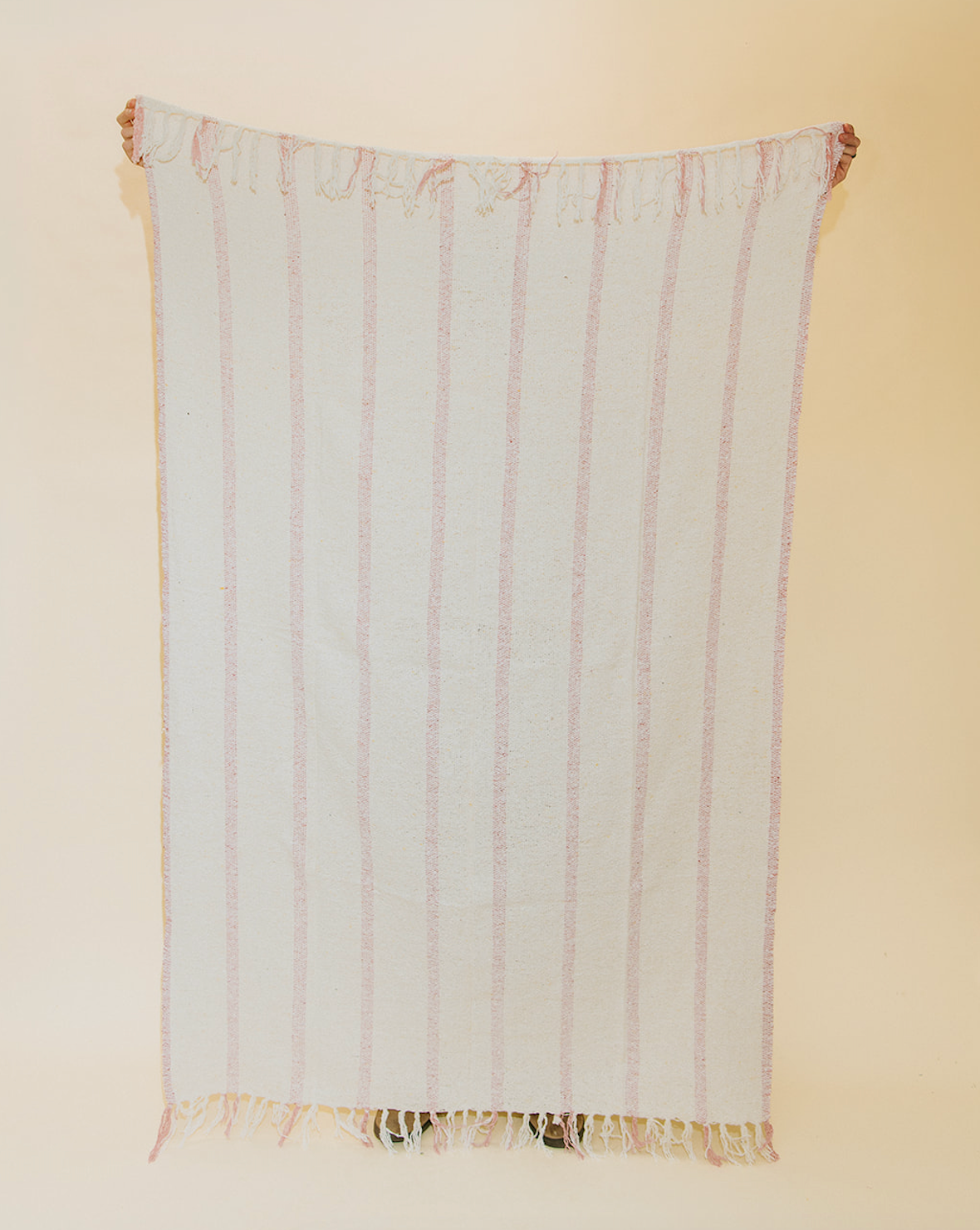 A rectangular woven textile hanging flat against a plain beige wall, featuring vertical pale pink stripes on an off-white base and fringed edges at the top and bottom.