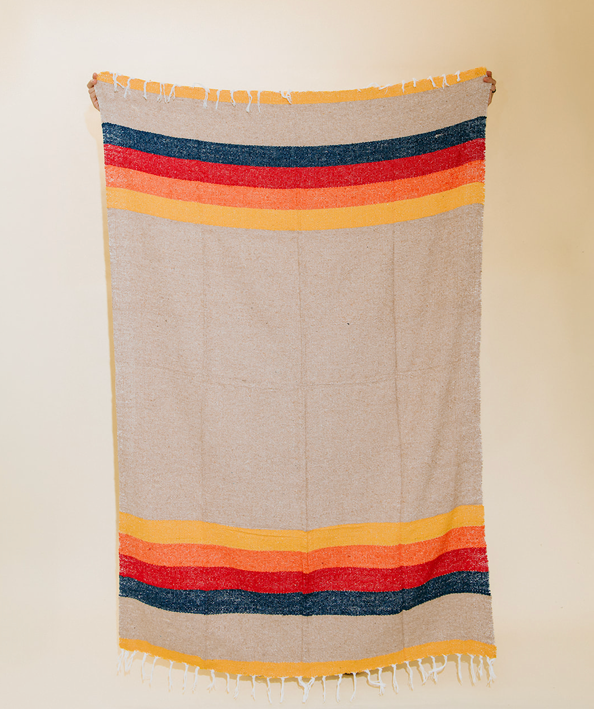 A woven Mexican-style blanket with horizontal stripes in navy blue, red, orange, and yellow on a light beige background, featuring white fringe on the top and bottom edges.