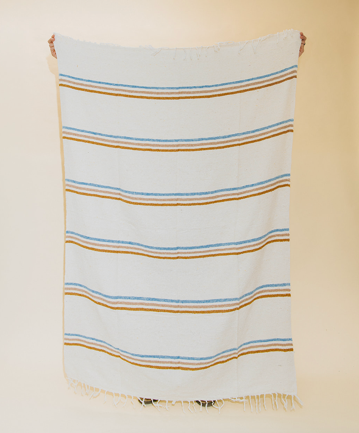 A white woven throw blanket with horizontal stripes in light blue and mustard brown, featuring fringed edges, hanging against a plain beige background.
